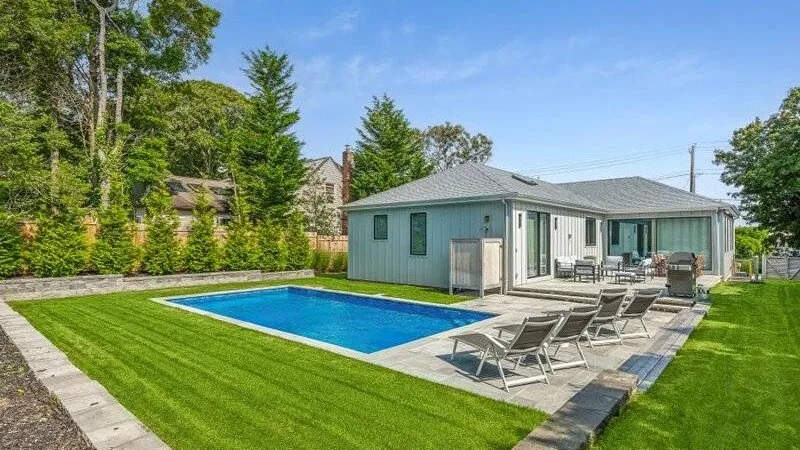 A swimming pool and lounge chairs sit beside a modern house on a green lawn.