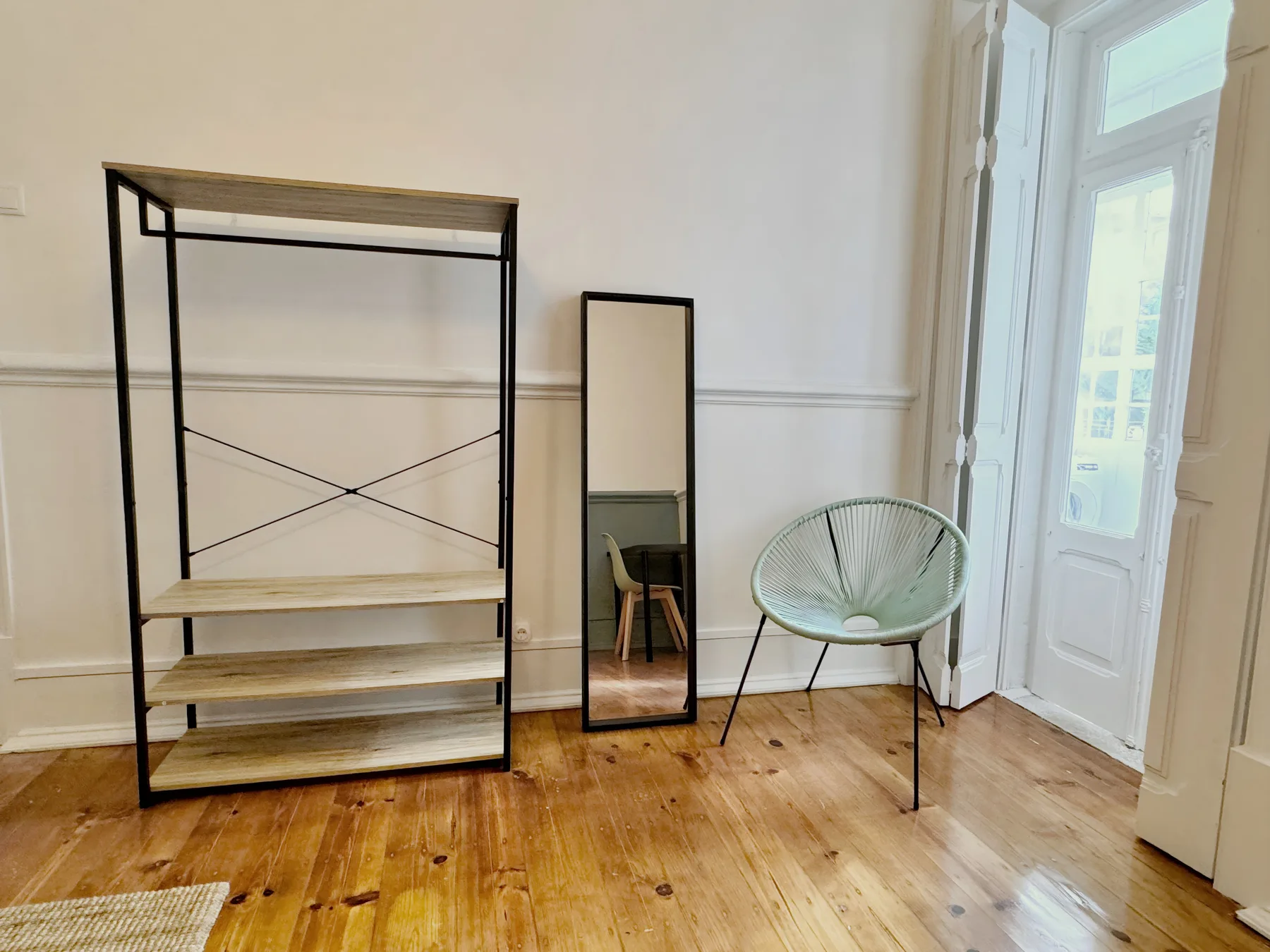 An airy room with an open shelving unit, a mirror, and a chair beside tall windows.