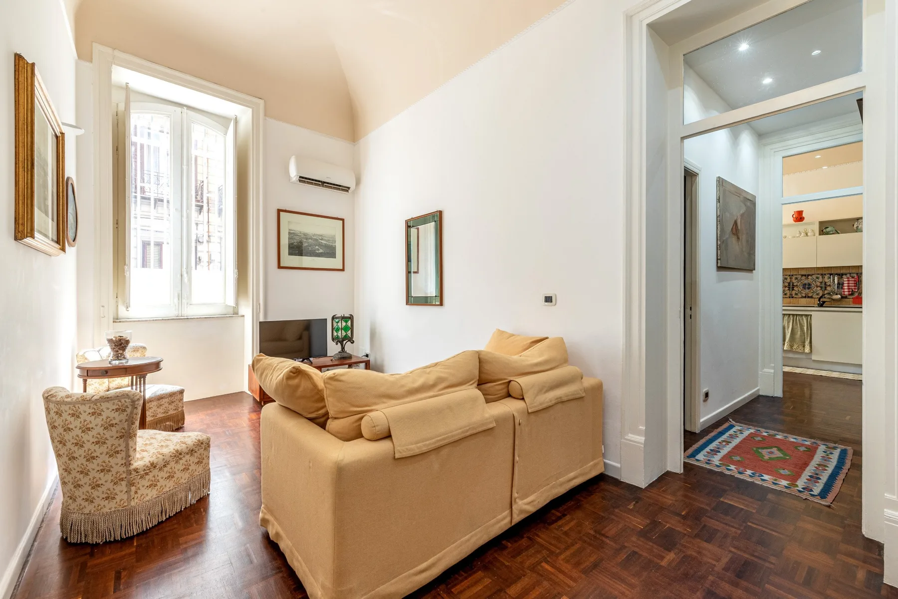 Una sala de estar con asientos color canela, una ventana y una puerta abierta a la cocina.
