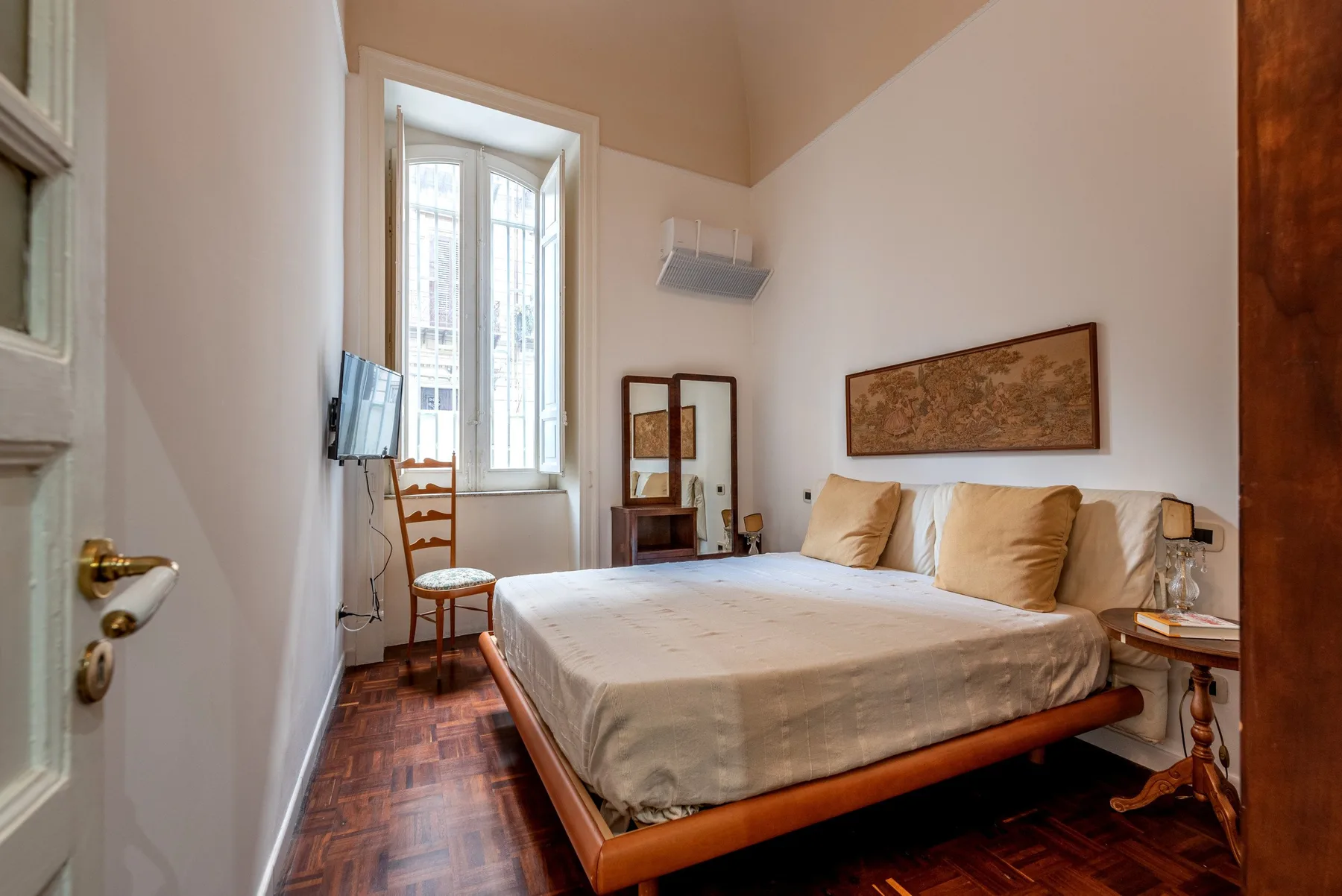 Un dormitorio con una cama, una ventana alta y un televisor pequeño.