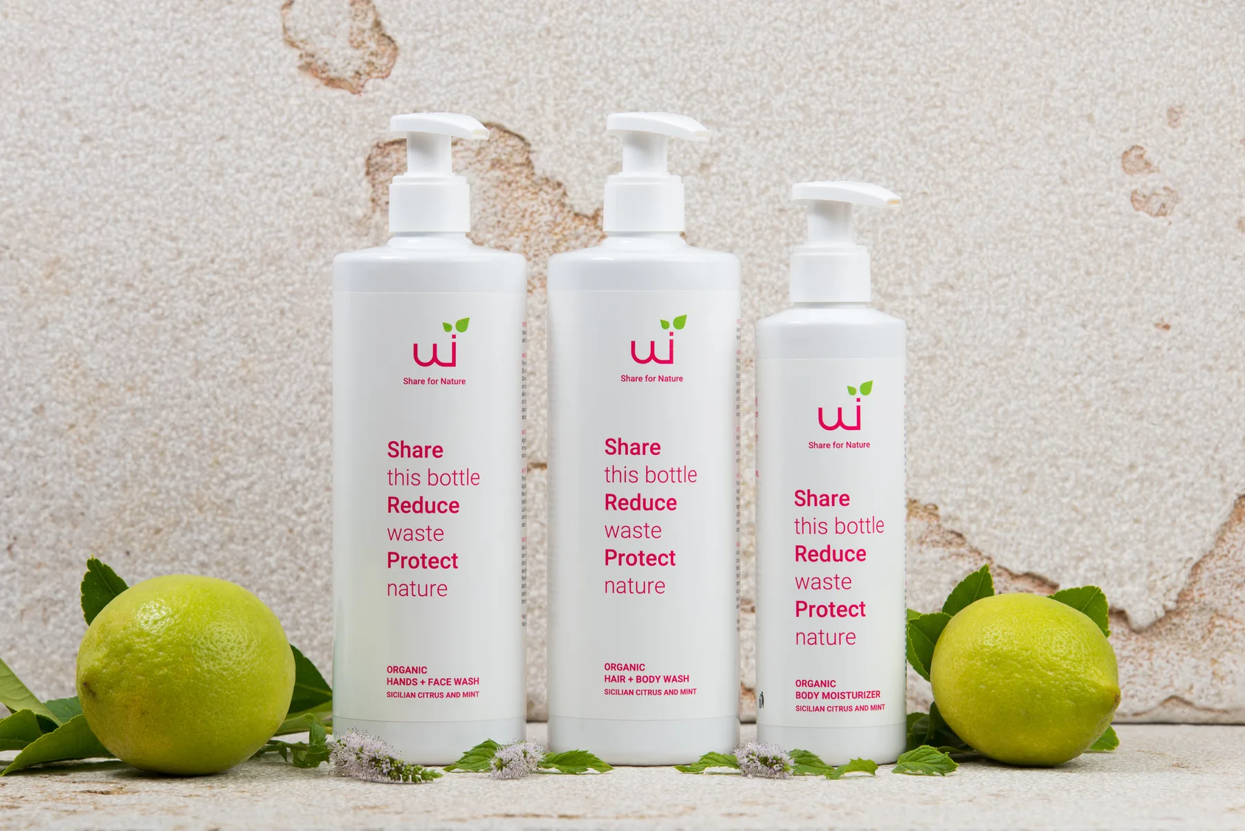 Tres botellas blancas con dispensador están entre cítricos verdes frente a una pared texturizada.
