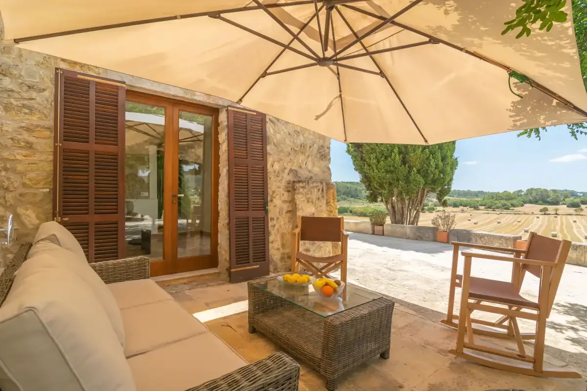 A shaded patio with outdoor seating and a table beside a stone house.