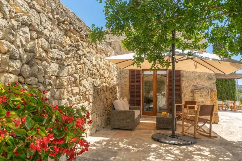 A stone patio has outdoor seating beneath a large shade umbrella and leafy tree.