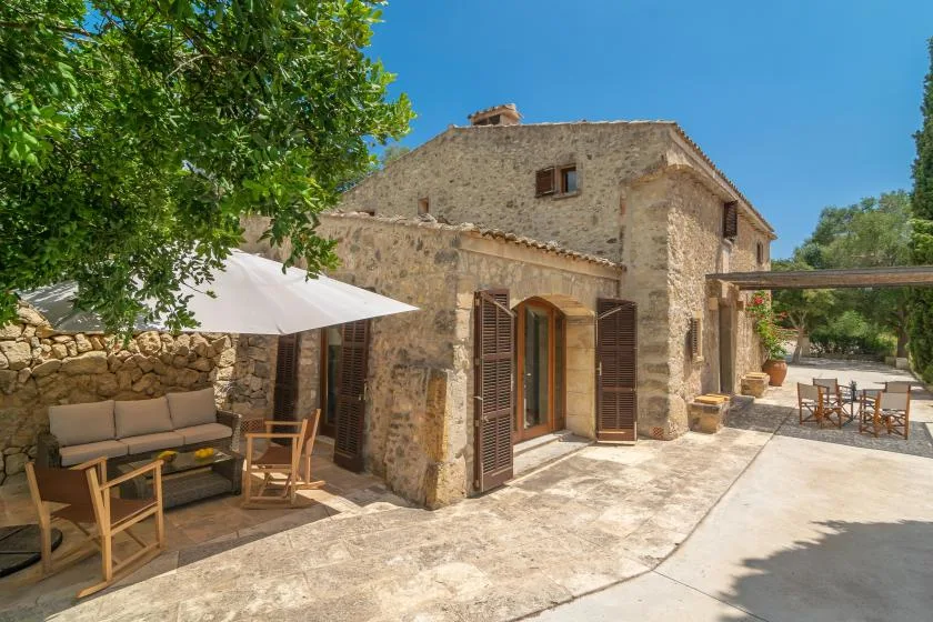 A stone house opens onto a sunlit patio with outdoor seating and an umbrella.