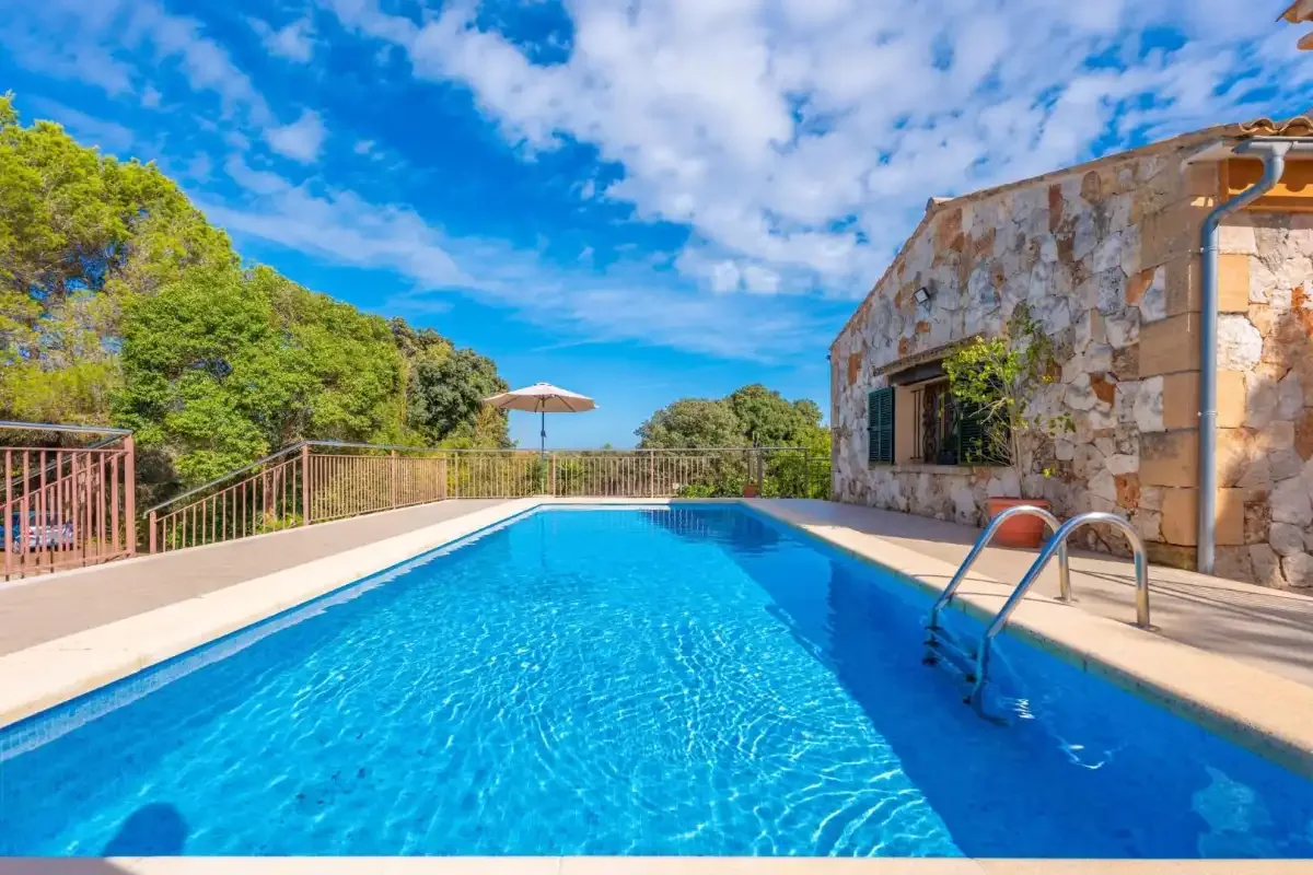 Una piscina junto a un edificio de piedra, con árboles y cielo azul.
