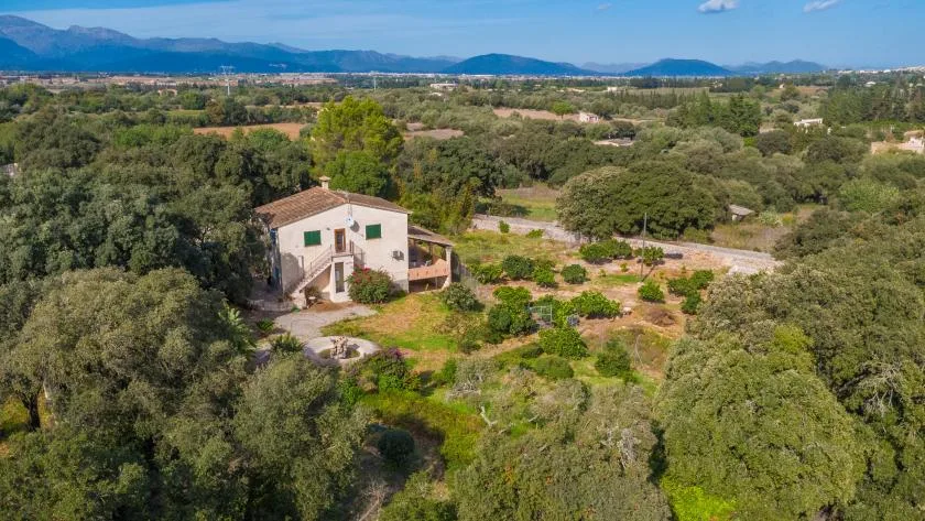 Una casa rural está entre árboles y campos cultivados, con montañas a lo lejos.