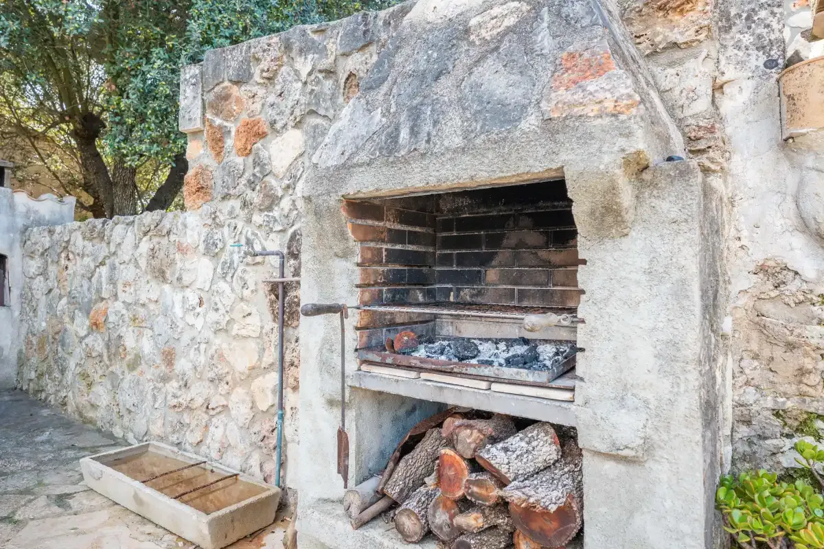 Una chimenea exterior de piedra está junto a un muro bajo y árboles.