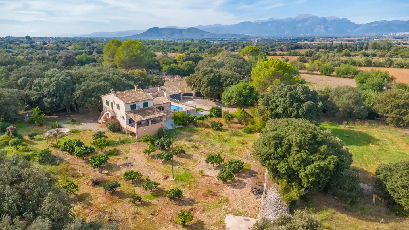 Una casa rural se encuentra entre árboles y campos, con montañas a lo lejos.