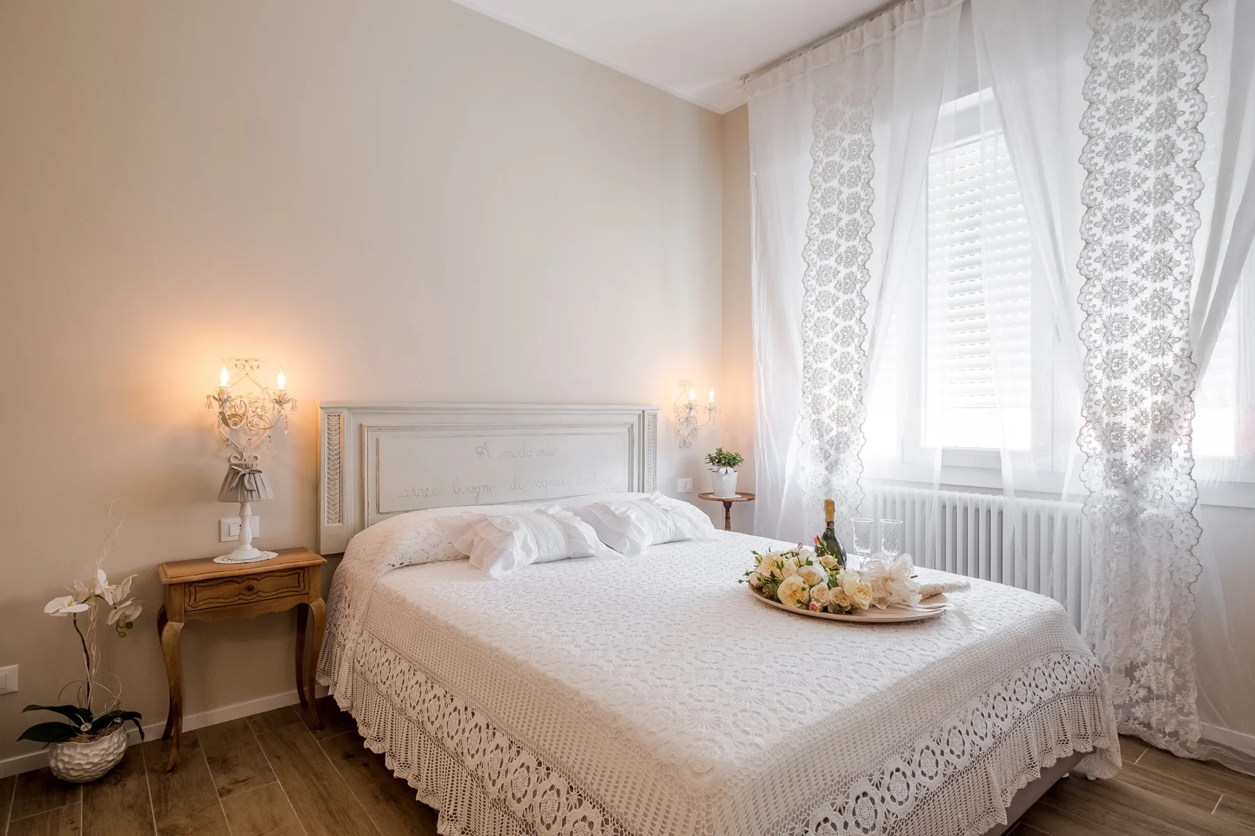 A bright bedroom with a lace-covered bed beside a large curtained window.