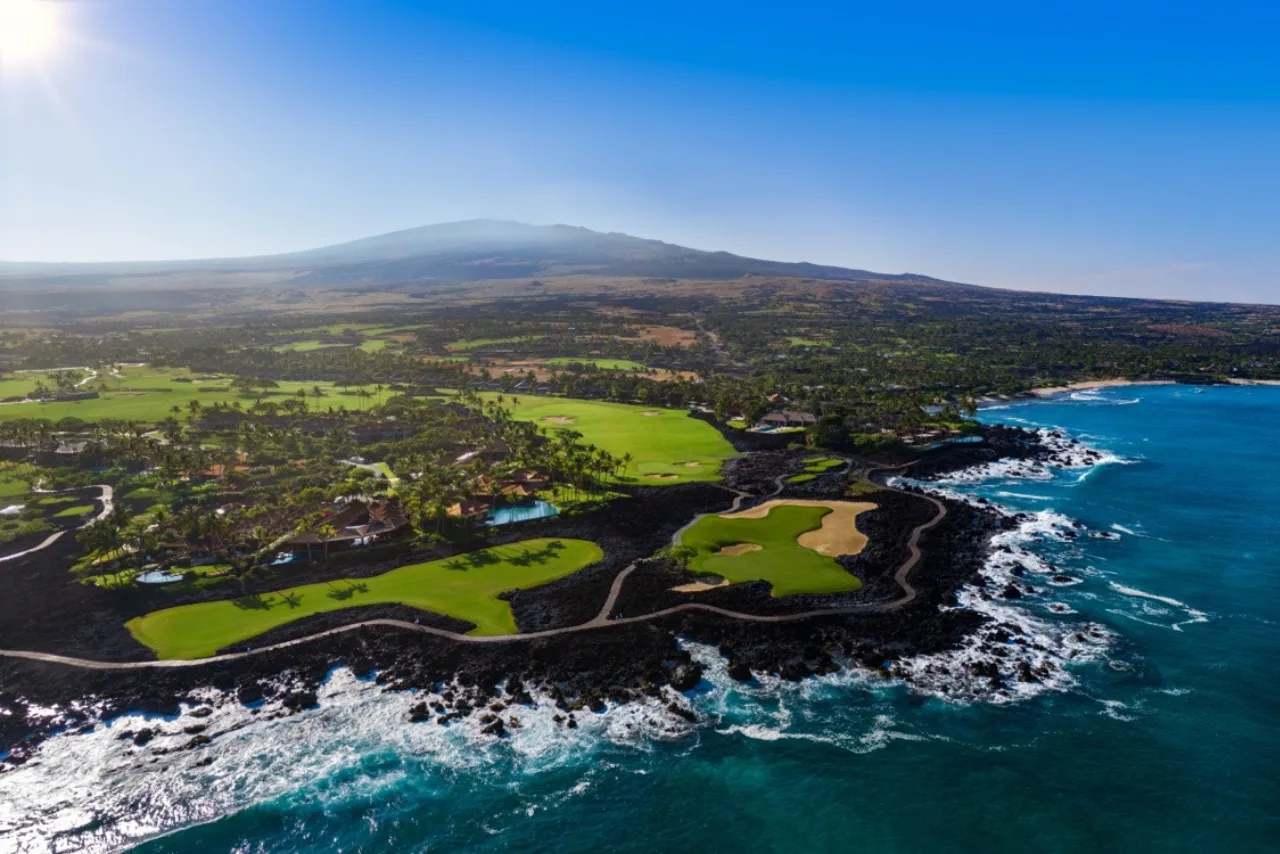 Paisaje costero aéreo con campos verdes, costa rocosa oscura y olas del océano.