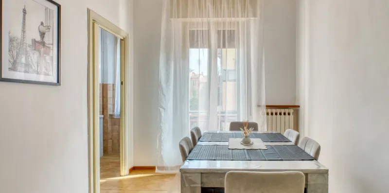 A dining area with a table and chairs beside a tall curtained window.