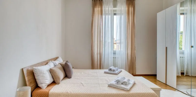 A bedroom with a neatly made bed and a tall curtained window.
