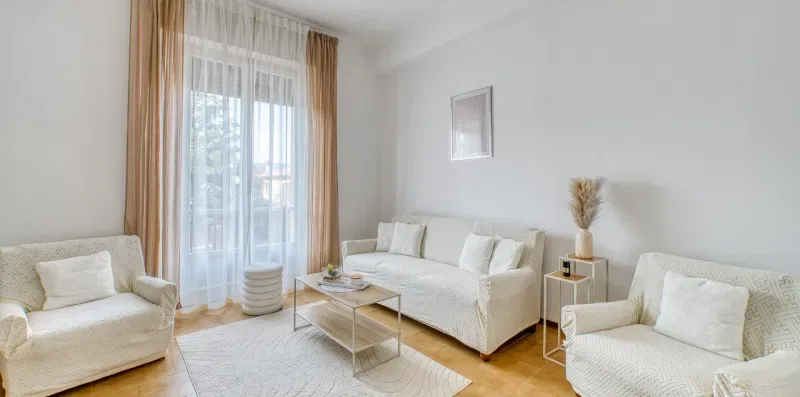 A bright living room with light-colored seating and a large curtained window.
