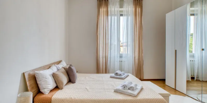 A bedroom with a neatly made bed beside a tall curtained window.