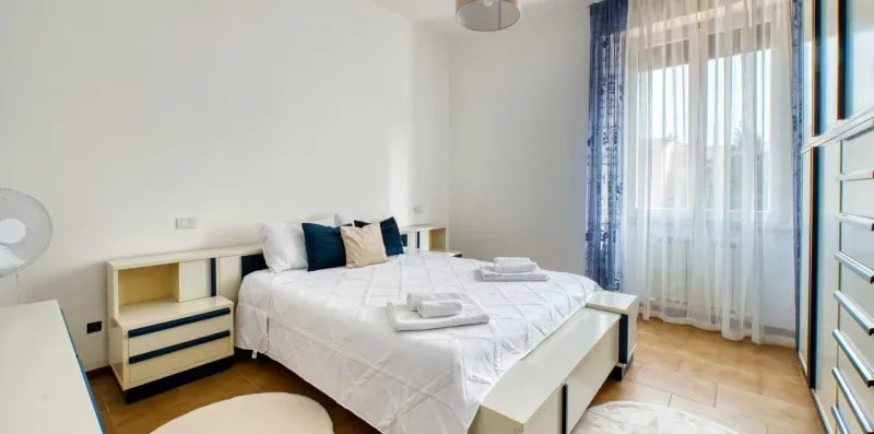 A bright bedroom with a neatly made bed and a large curtained window.