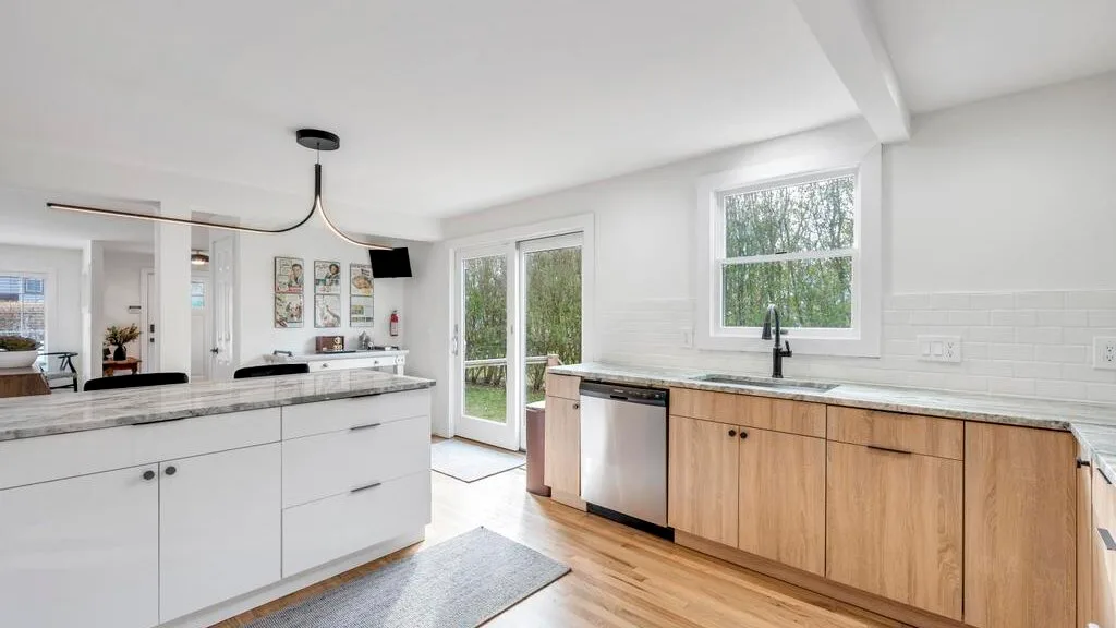 Una cocina luminosa con encimeras blancas, gabinetes de madera y ventanas con vistas a la vegetación.