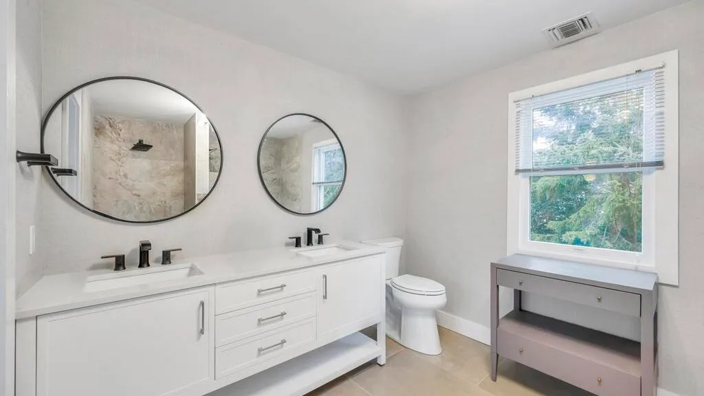 Un baño luminoso tiene un lavabo doble, espejos y un inodoro junto a una ventana.