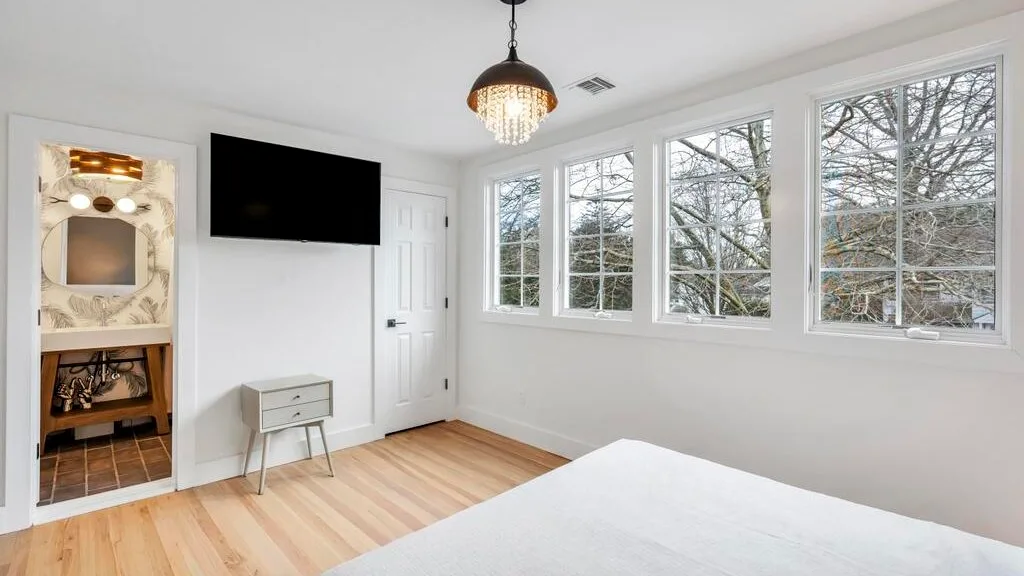 Un dormitorio luminoso con una cama, un televisor en la pared y ventanas grandes.