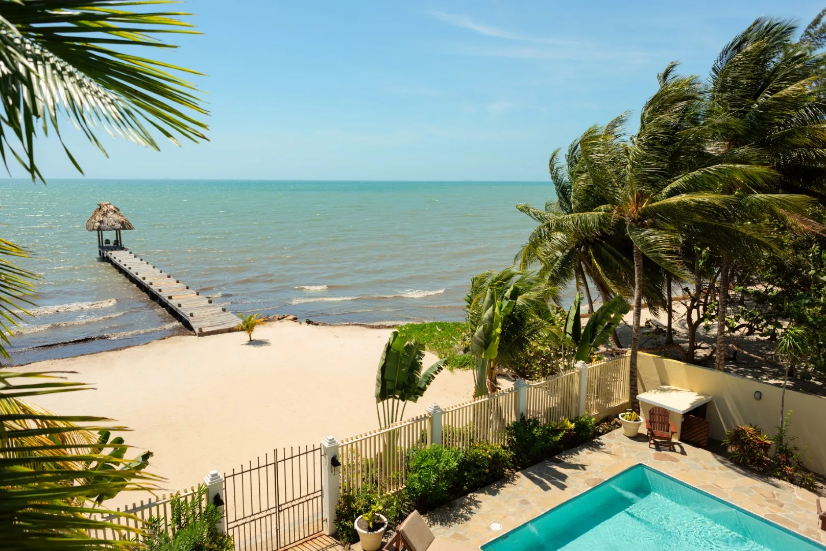A sandy beach and ocean with a pier, palm trees, and a swimming pool.