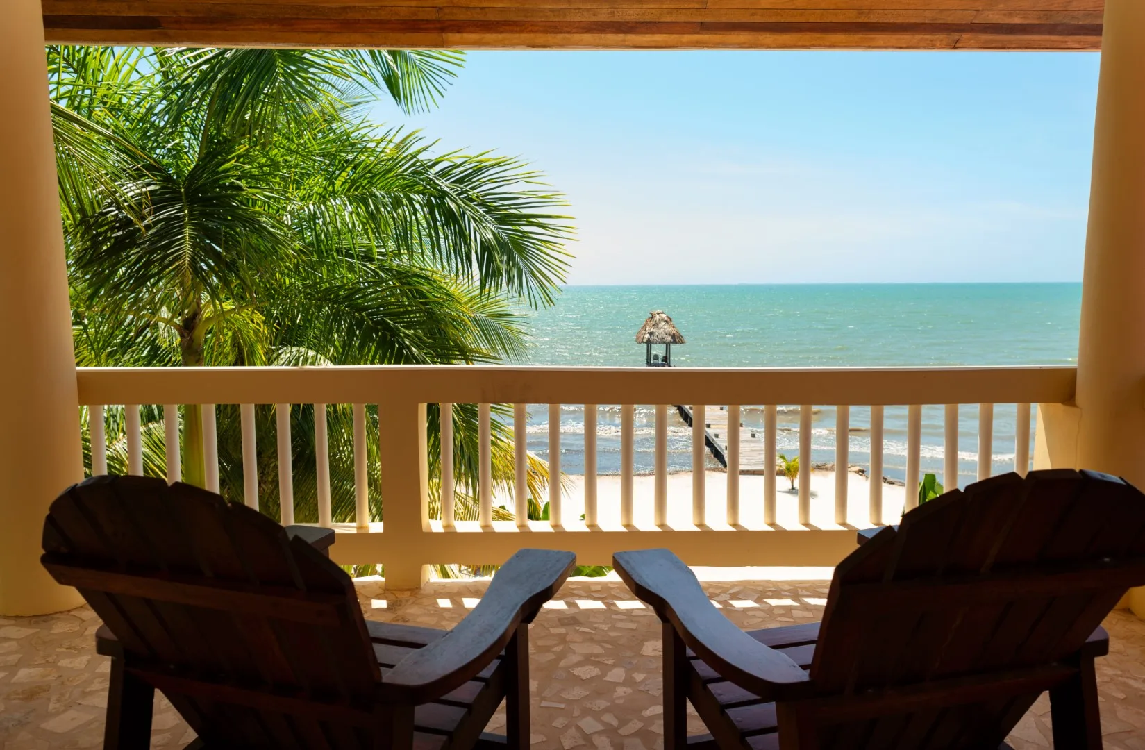 A covered balcony overlooks the beach and ocean, with two lounge chairs in the foreground.