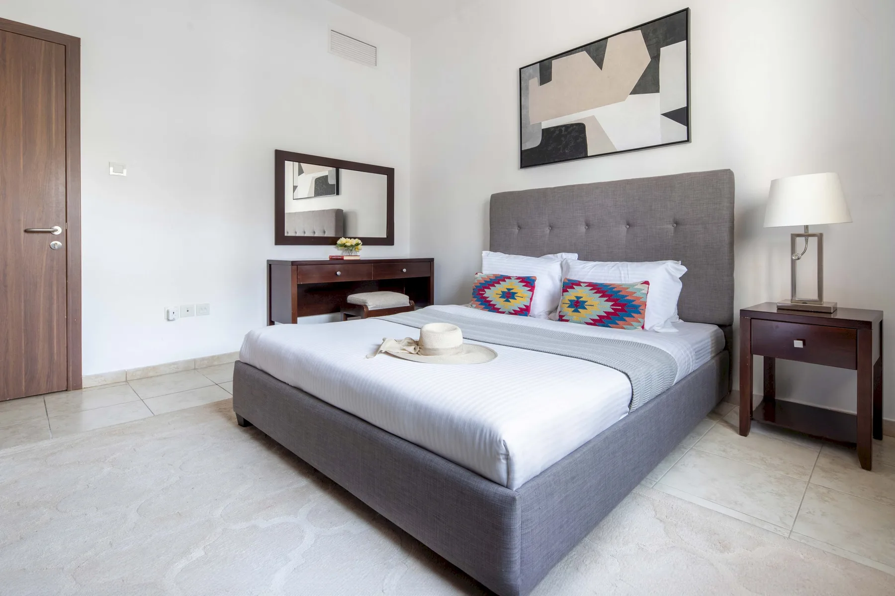 Un dormitorio con una cama gris, cojines estampados de colores y una mesita de noche.