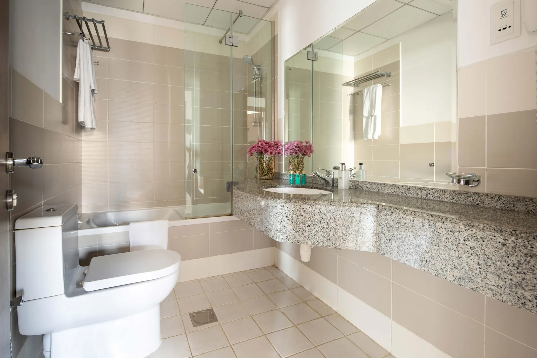 Un baño luminoso con azulejos, una ducha de vidrio y un largo lavabo.