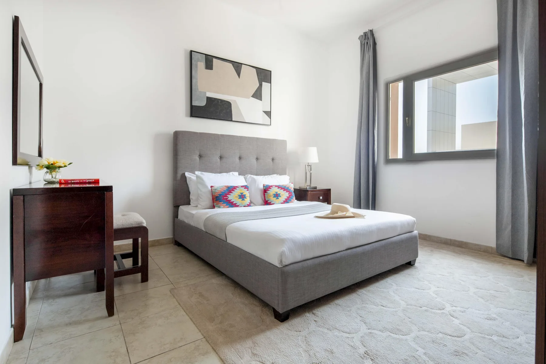 Un dormitorio con una cama tapizada gris, cojines coloridos y una ventana grande.
