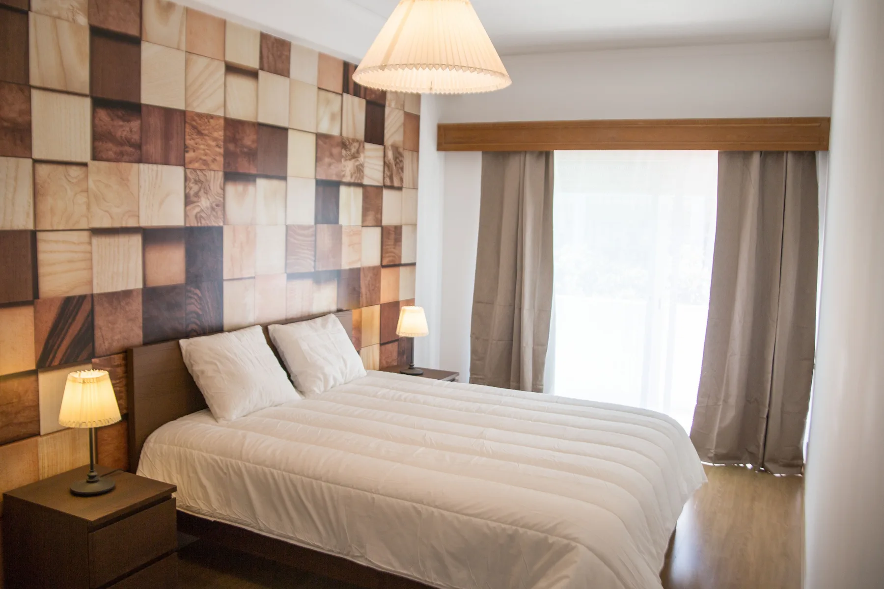 Un dormitorio con una cama doble, mesitas de noche y una ventana luminosa con cortinas.