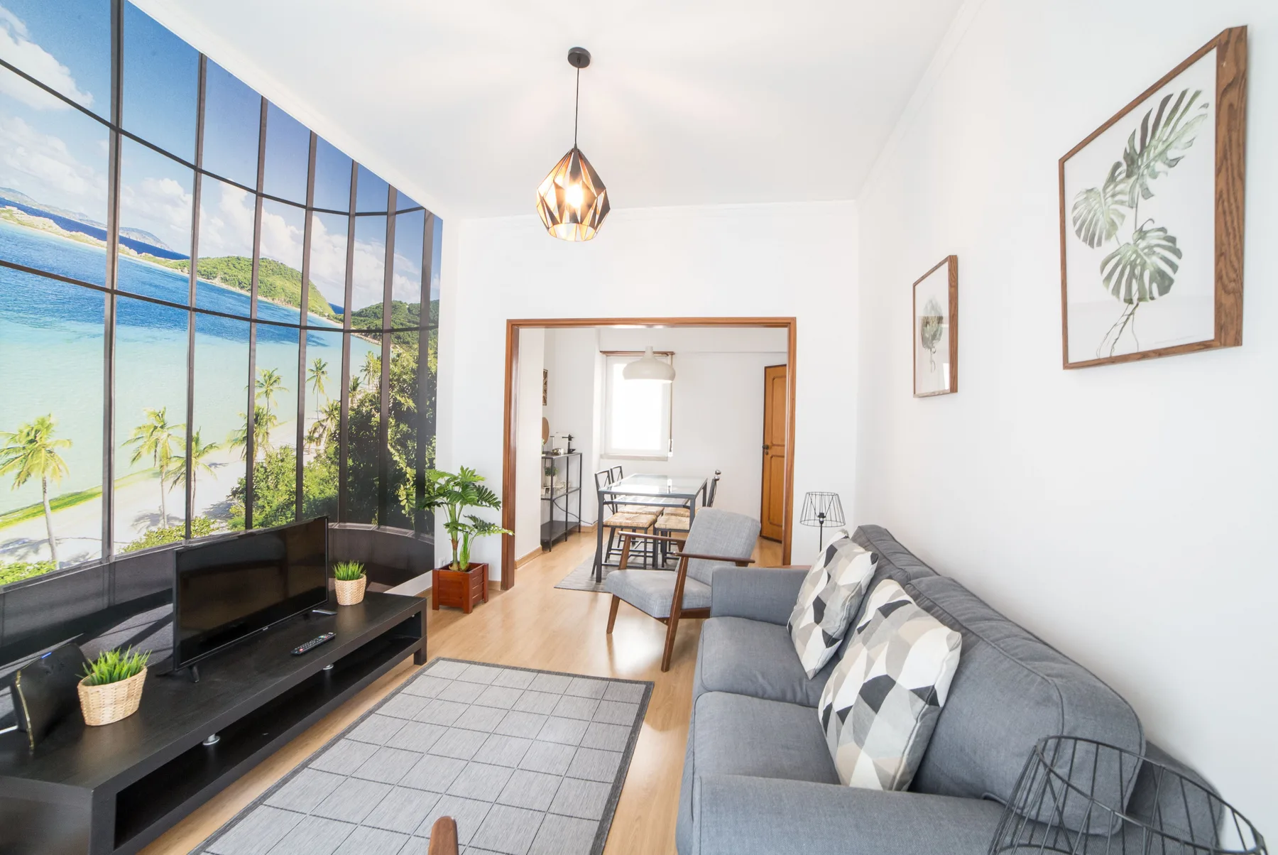 Una sala luminosa con asientos grises, un televisor y un gran mural tropical.