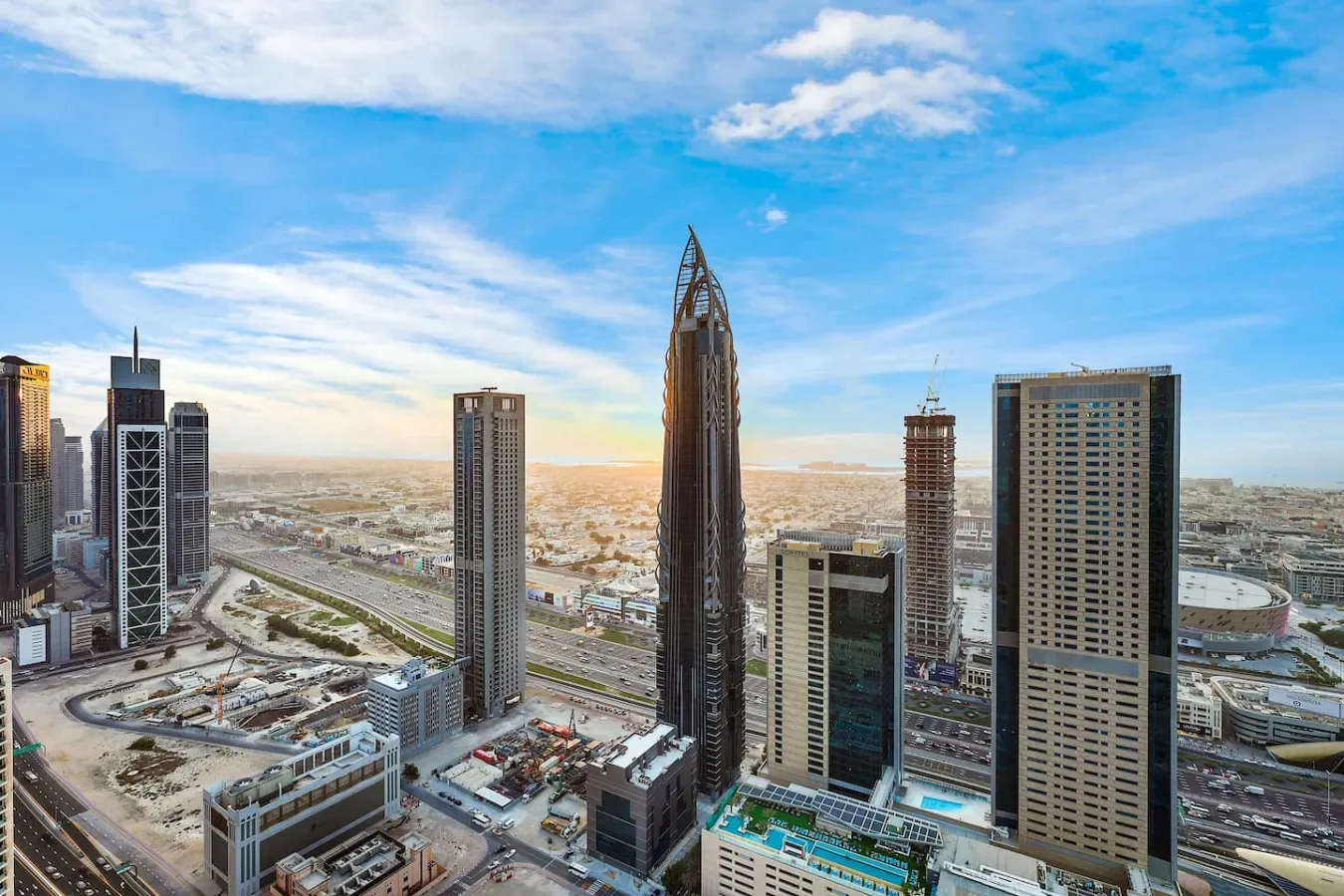 A city skyline of modern high-rise buildings beneath a blue, cloud-filled sky.