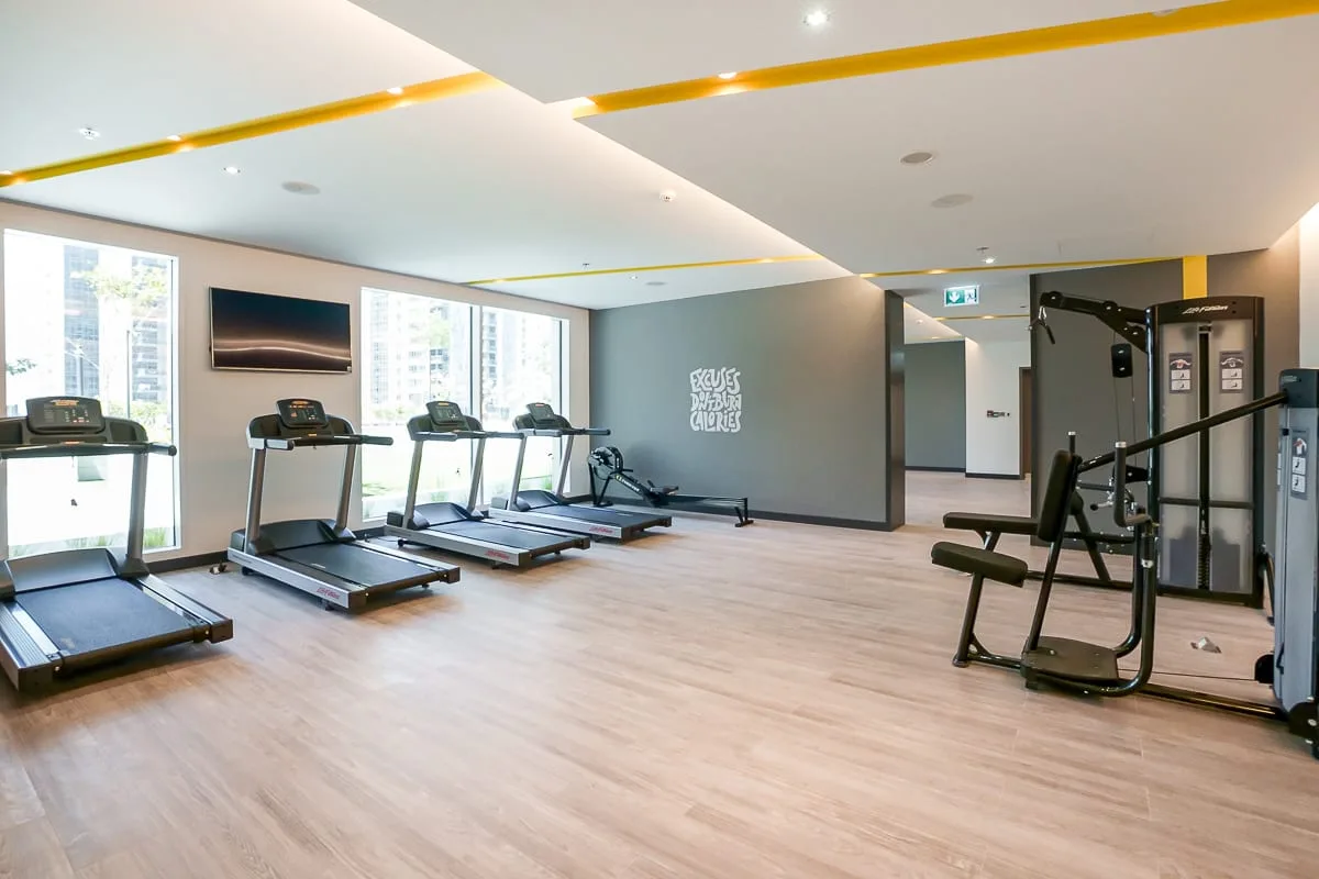 Un gimnasio luminoso con cintas de correr y equipos de ejercicio junto a las ventanas.
