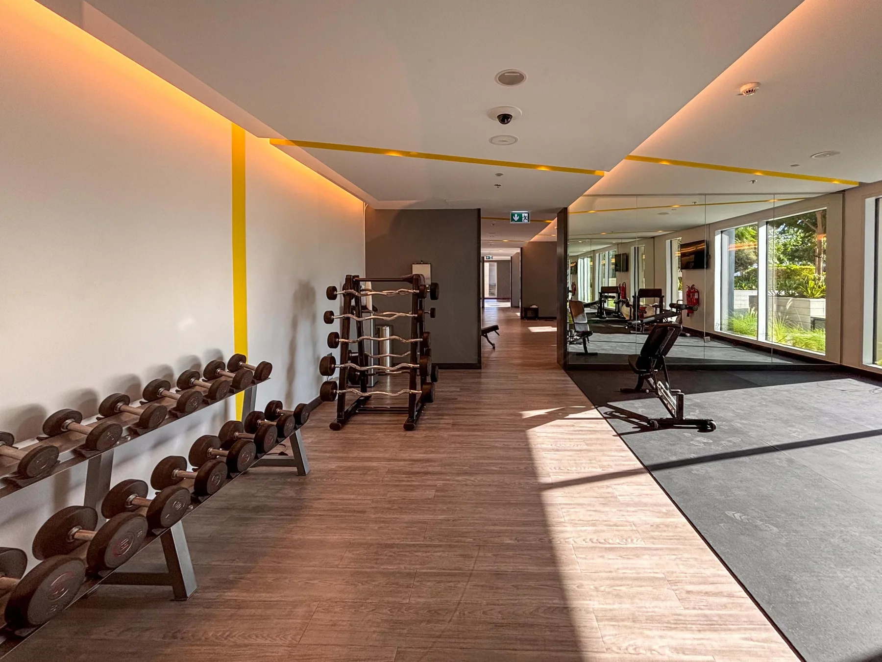Un gimnasio con soportes para mancuernas, equipo de ejercicio y grandes ventanas.