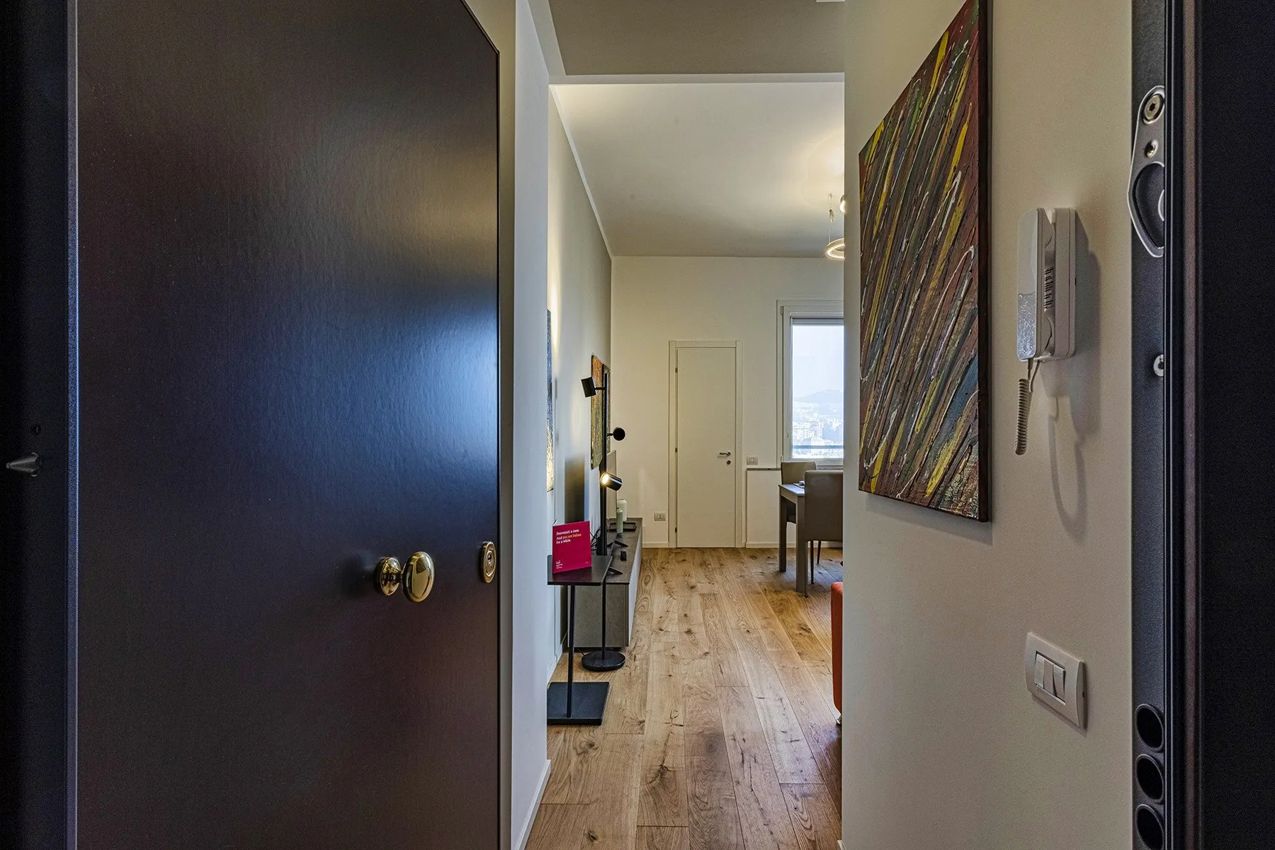 An apartment entryway opens onto a hallway with wood flooring and a wall-mounted intercom.