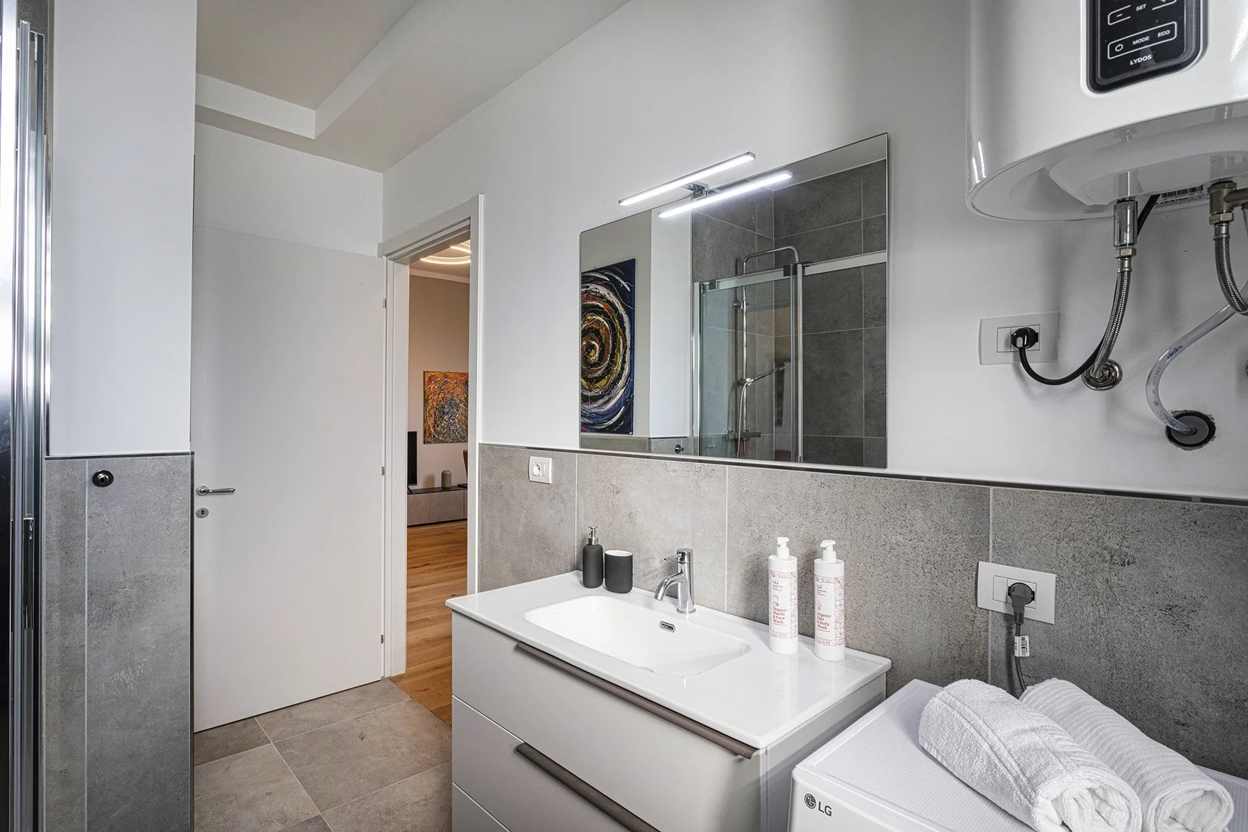 A bathroom vanity with a sink, mirror, and toiletries beside a washing machine.