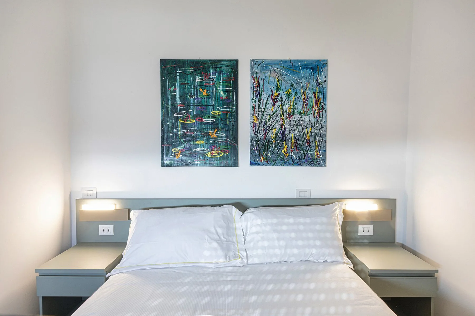 A bedroom with a neatly made bed and two abstract paintings above the headboard.