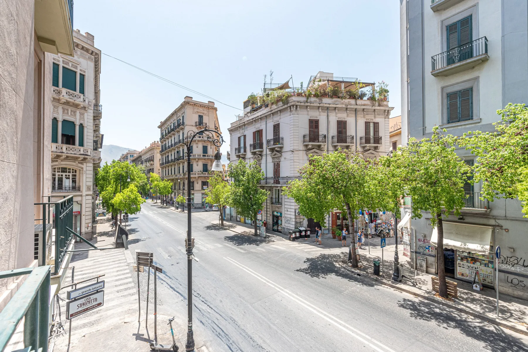 A sunlit city street lined with multistory buildings and leafy trees.