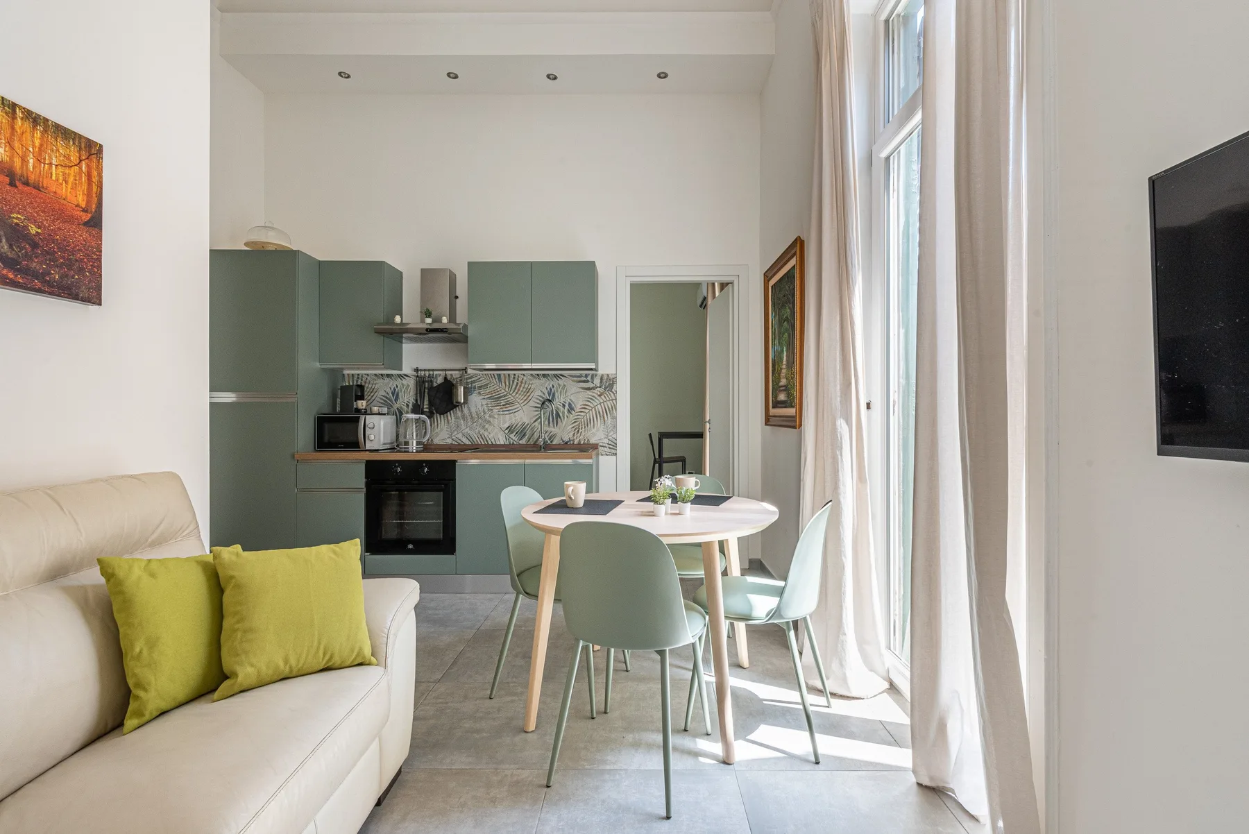 A bright living and dining area with green kitchen cabinets and tall windows.
