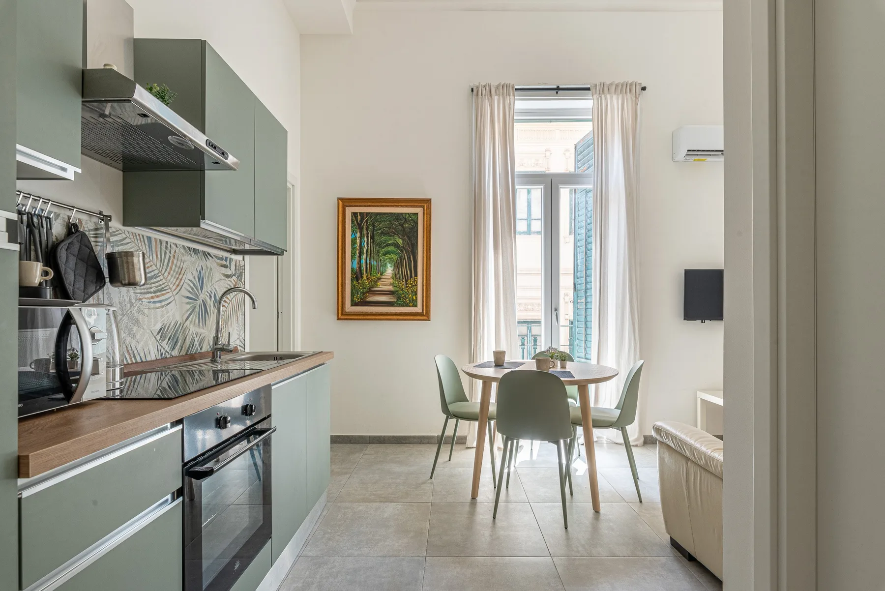A modern kitchen opens into a dining area with a table beside tall windows.