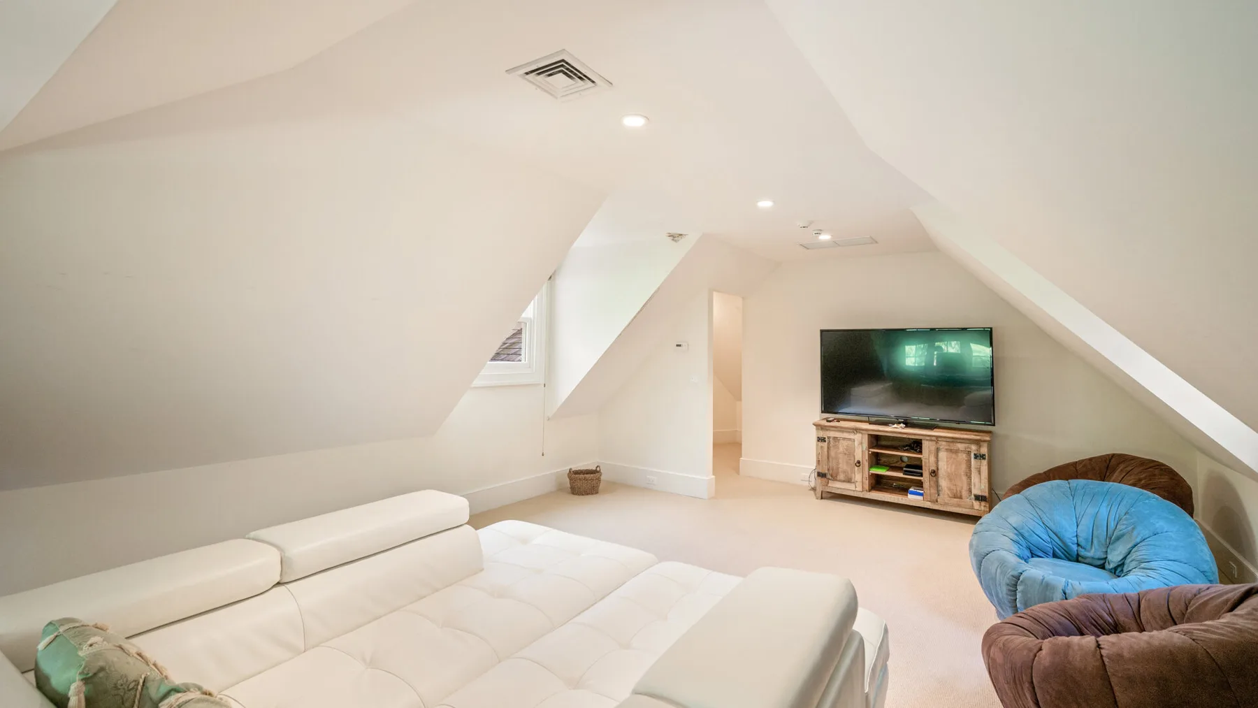 Una luminosa sala de estar en el ático, con techos inclinados bajos y asientos frente a un televisor.