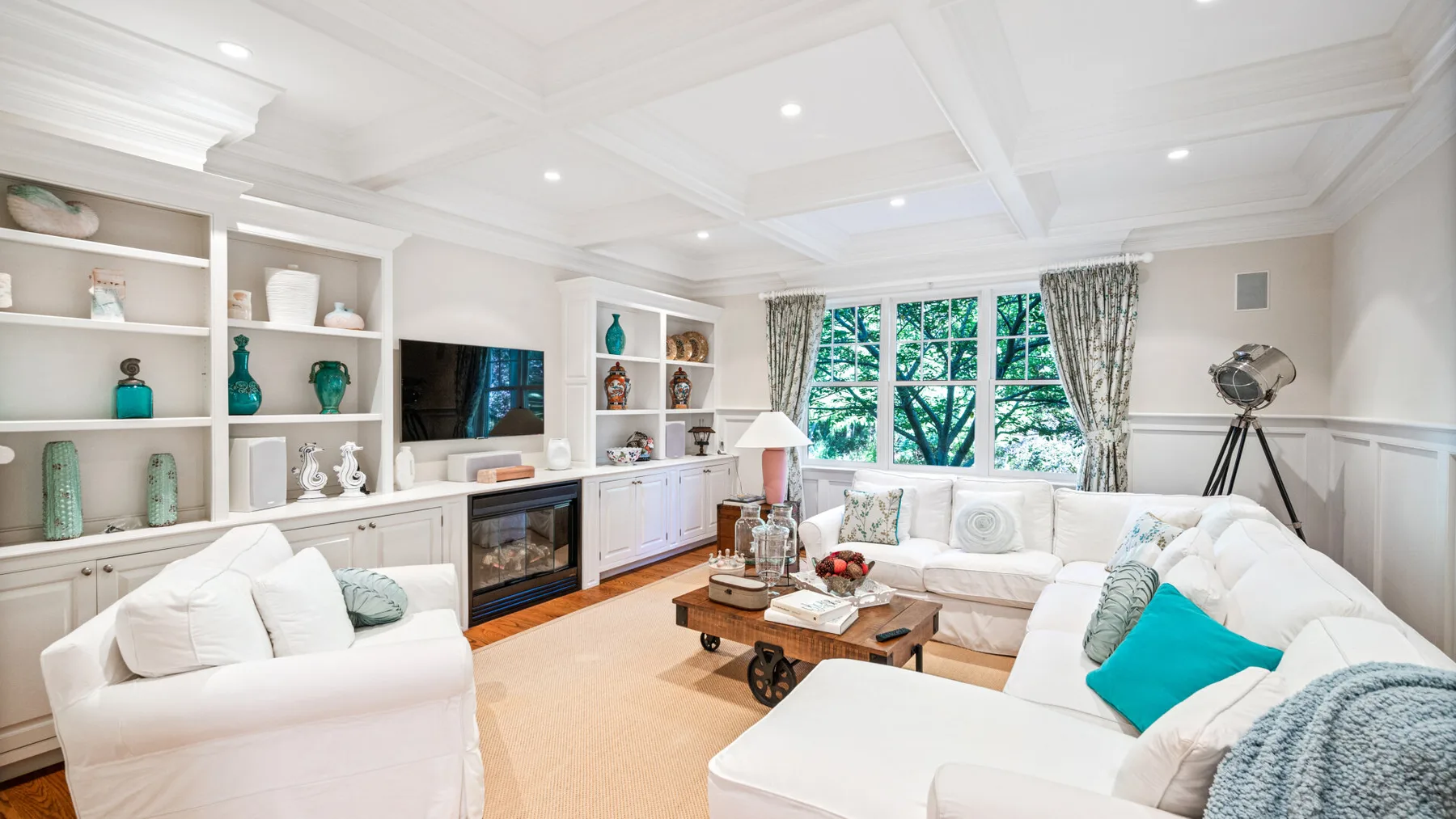 Una sala luminosa con asientos blancos, estantes empotrados y grandes ventanas con cortinas.