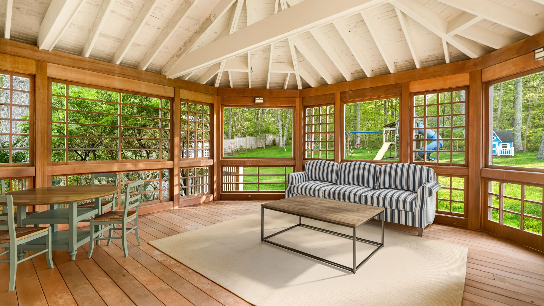 A bright sunroom with a striped sofa, dining table, and windows overlooking greenery.