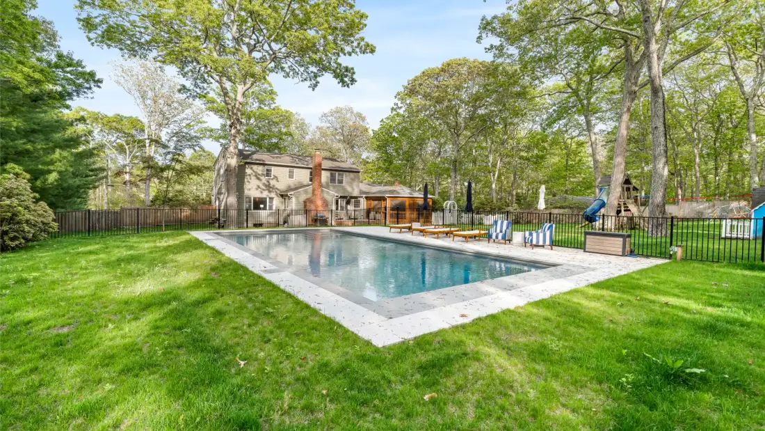 Una piscina en el patio está junto a una casa, rodeada de césped y árboles.