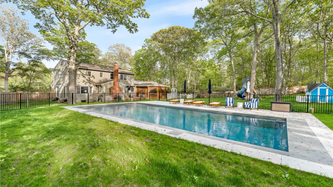 An outdoor swimming pool sits beside a lawn, with trees and seating nearby.