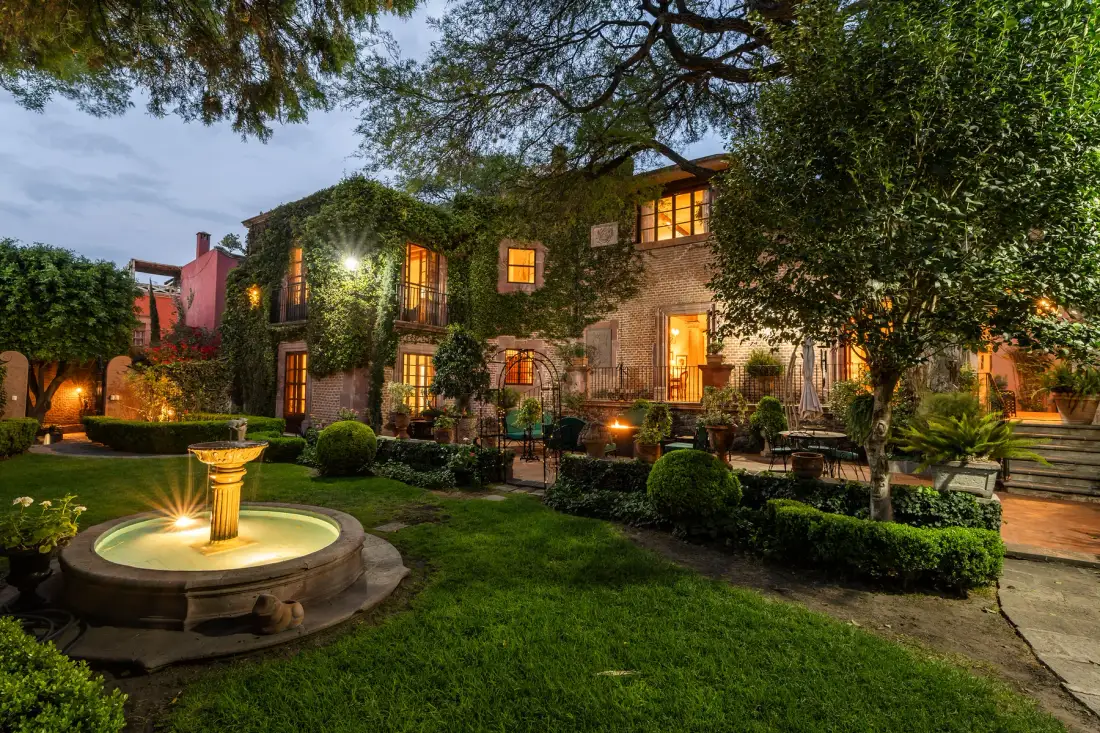 An ivy-covered house overlooks a landscaped garden with a lit fountain.