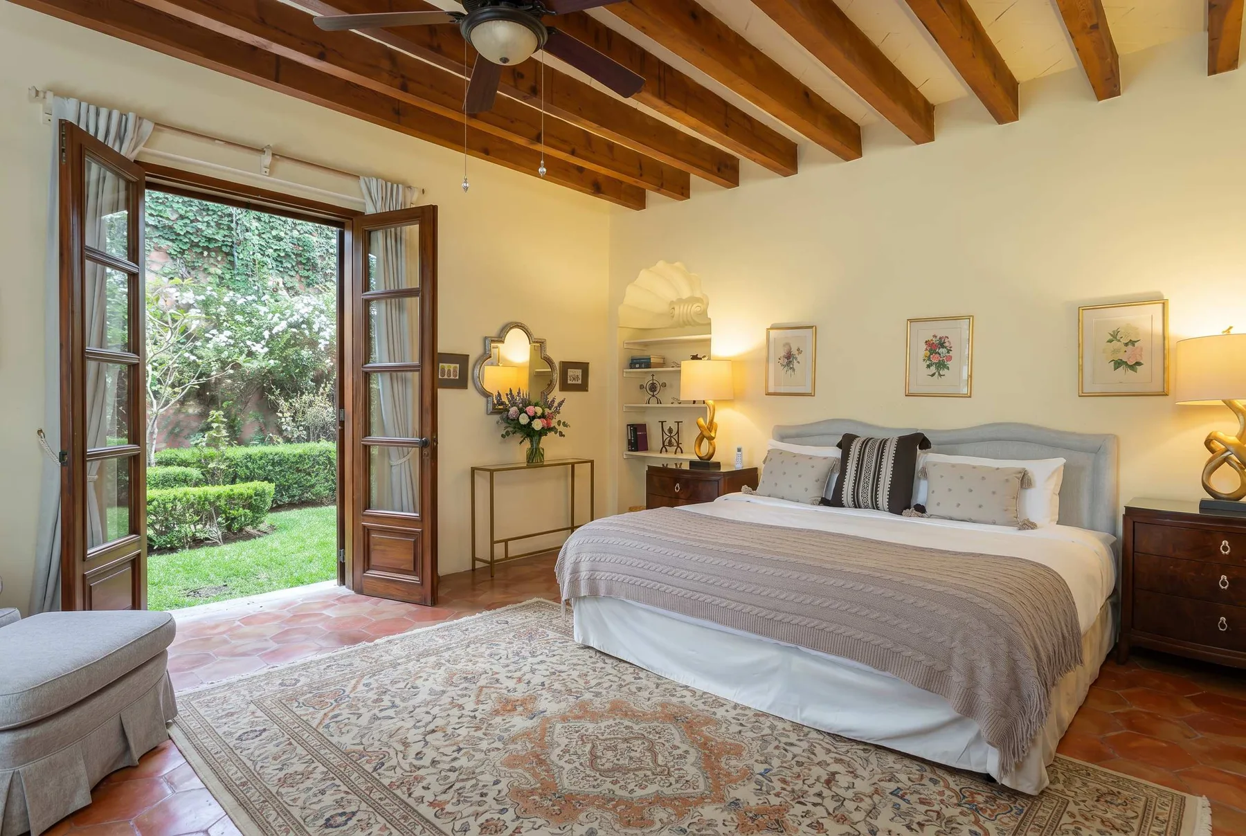 A bedroom with a large bed, patterned rug, and open doors overlooking a garden.