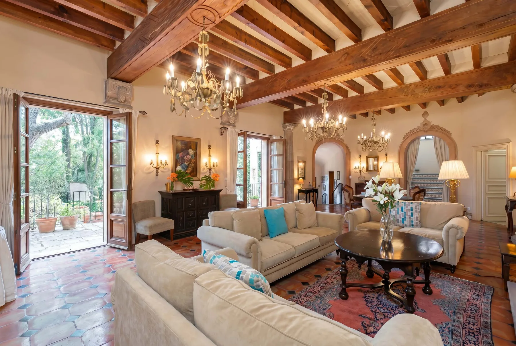 A spacious living room with seating, chandeliers, and exposed wooden ceiling beams.