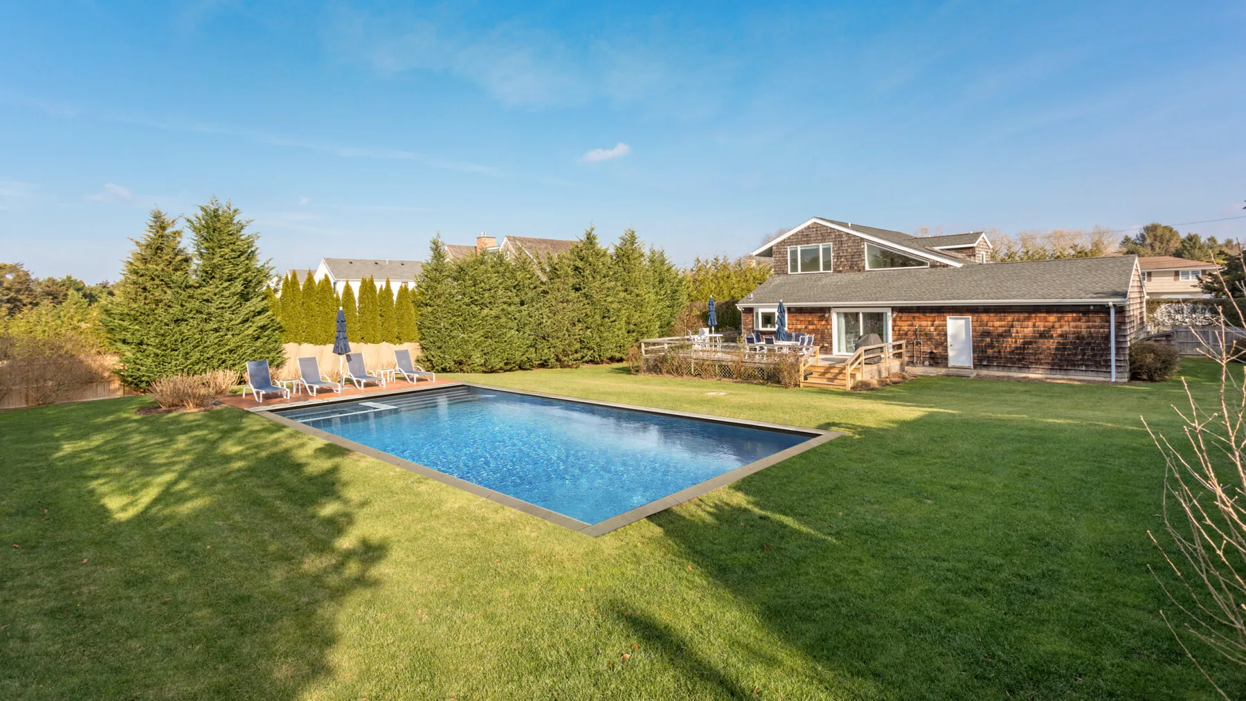 Un césped trasero rodea una piscina rectangular junto a una casa baja.