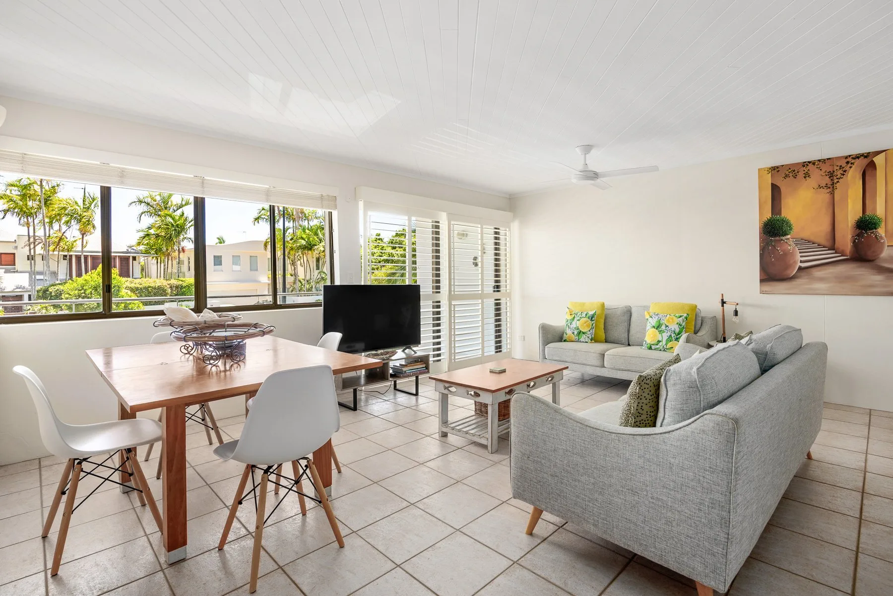 A bright living and dining area with gray seating, a television, and large windows.