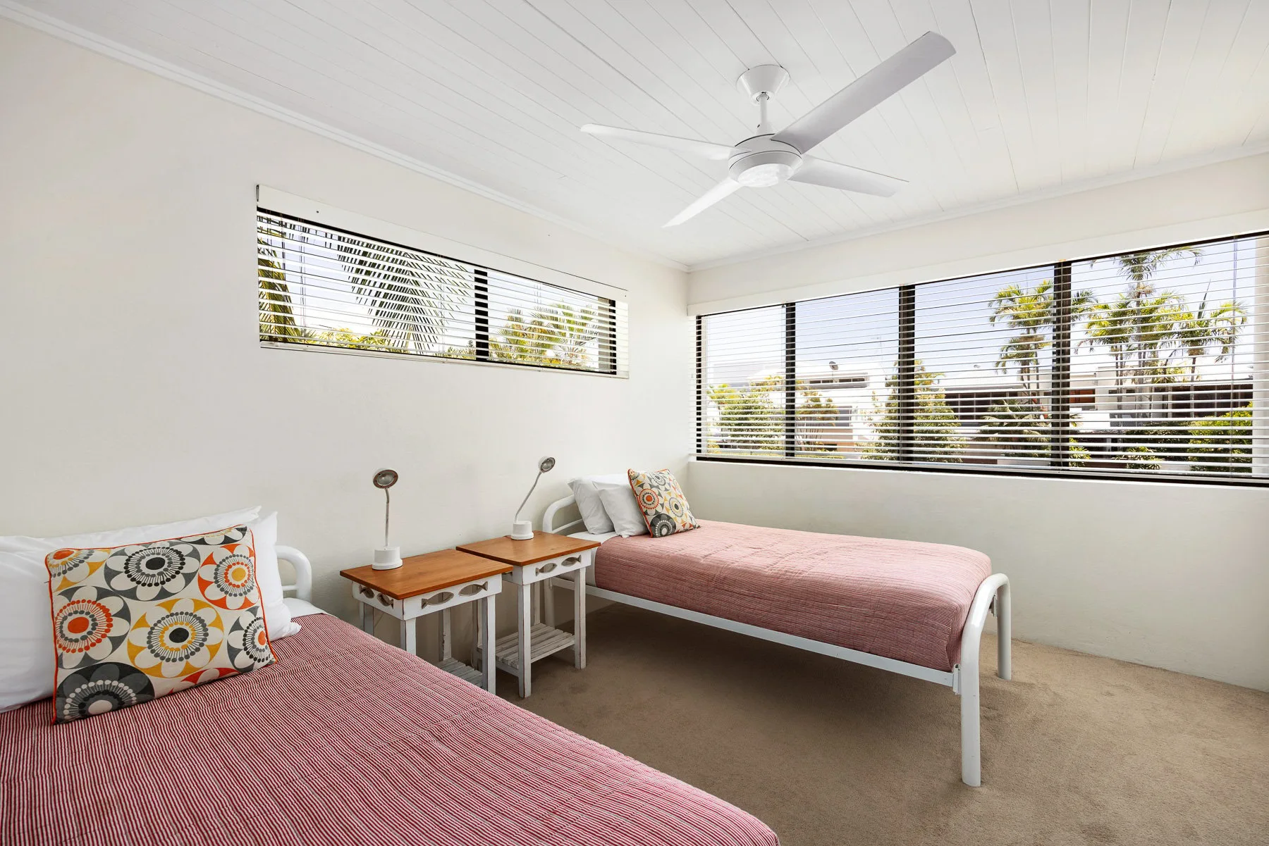 A bright bedroom with two beds and wide windows fitted with blinds.