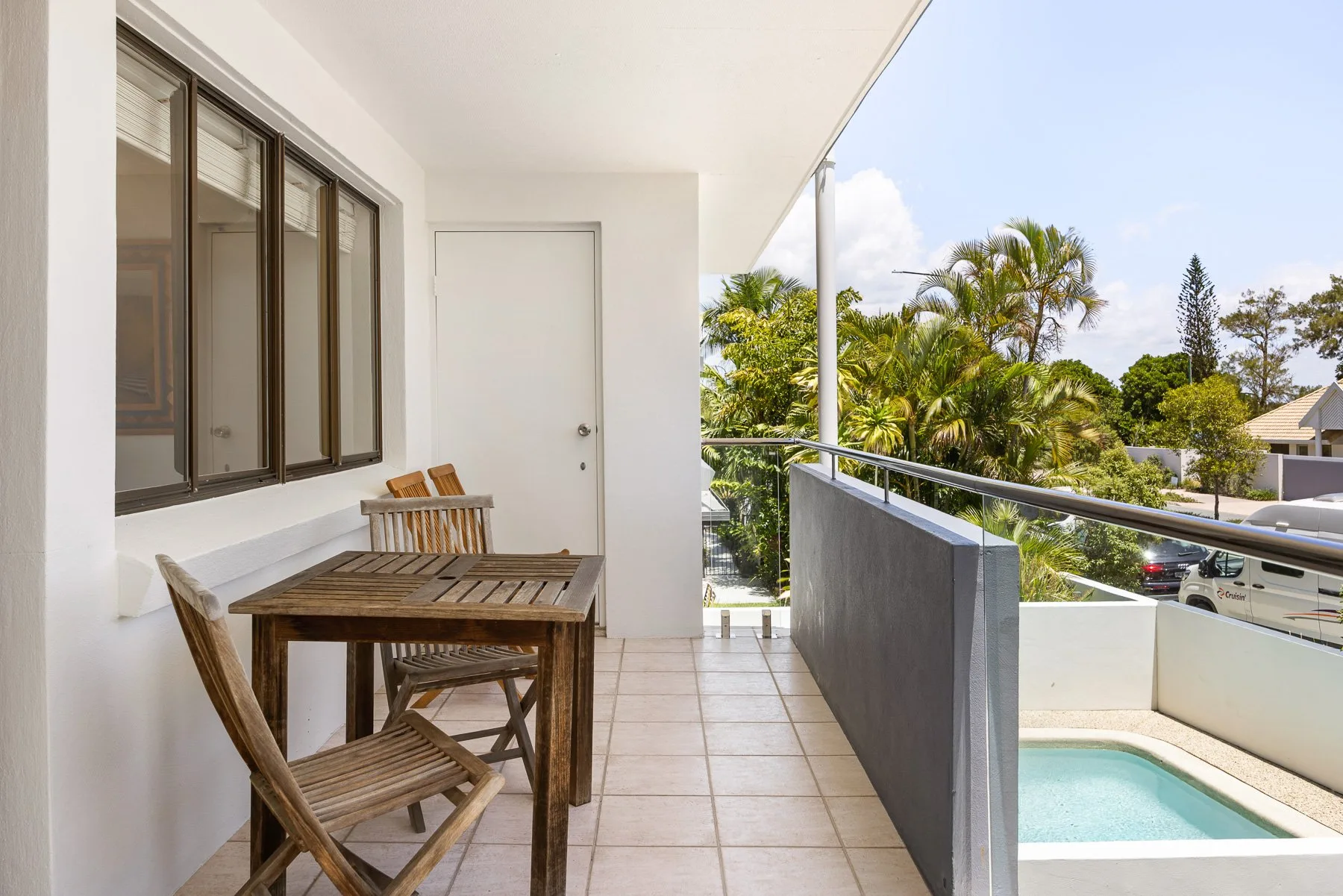 A covered balcony with a wooden dining table overlooks a swimming pool and palm trees.